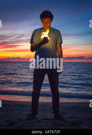 Un jeune homme se tient sur une plage au coucher du soleil et regarde son smartphone brûlant alors que les flammes illuminent son visage Banque D'Images