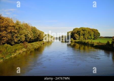 Une grande rivière coule calmement entre un paysage d'automne avec des arbres et des prairies avec des feuilles colorées, sous un ciel bleu. Banque D'Images