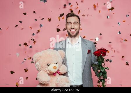 homme souriant en costume tenant un ours en peluche et des roses pour la saint valentin, sur le rose avec des confettis Banque D'Images