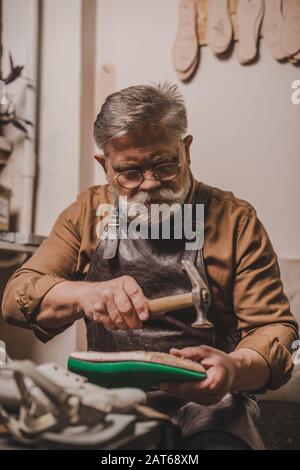 la chaussure pavée senior, barbu, répare la chaussure avec hummer en atelier Banque D'Images