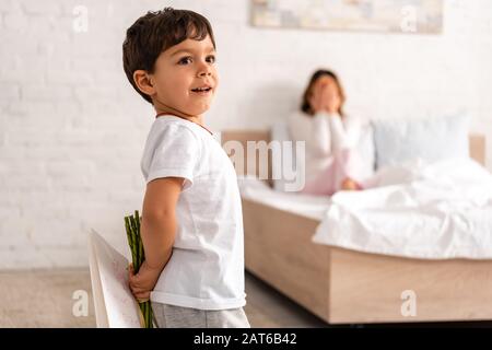 foyer sélectif d'adorable garçon tenant des fleurs et carte de fête des mères, et femme assise dans le lit et couvrant les yeux avec les mains Banque D'Images