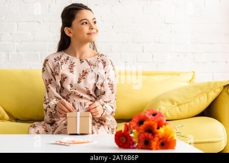 joli enfant souriant dans une élégante robe assise près de la table avec fleurs et boîte cadeau Banque D'Images