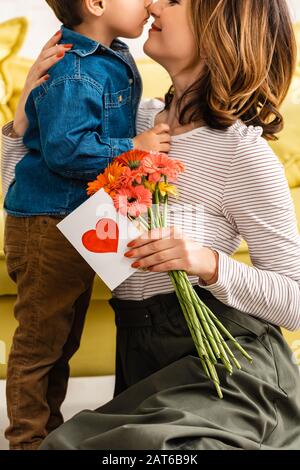 vue rognée d'une heureuse femme tenant des fleurs et carte de fête des mères avec le symbole du coeur tout en embrassant son adorable Banque D'Images