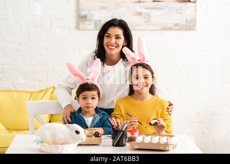 Heureuse femme et enfants dans des oreilles de lapin souriant à l'appareil photo tout en peignant des œufs de Pâques près de lapin blanc en osier Banque D'Images