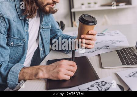 vue rognée de l'animateur barbu tenant la tasse en papier près des esquisses Banque D'Images