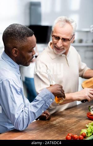 un homme africain américain qui tient de la bière et son ami souriant cuisinant dans la cuisine Banque D'Images