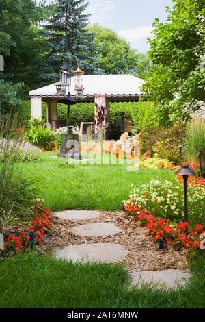 Sentier gris en pierre de pavillon à travers les bordures de paillis planté avec Miscanthus - plantes ornementales d'herbe, fleurs rouges de Begonia et belvédère dans le jardin d'arrière-cour Banque D'Images