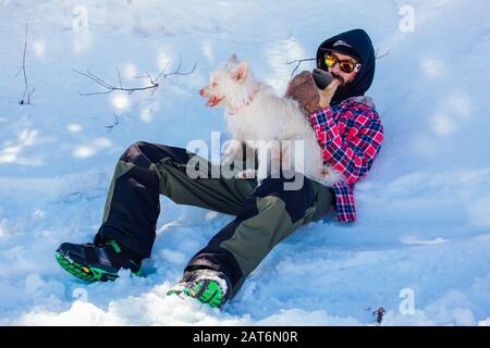 Sheregesh, Moscow, Russie - 08 Avril 2019 : Un homme se trouve dans un banc de neige et un chien blanc est assis sur elle Banque D'Images