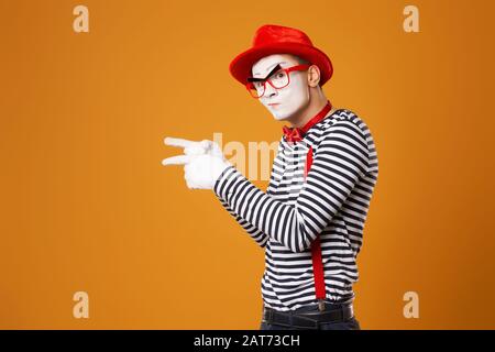 MIME dans Red Hat et dans la veste pointant avec sa main à côté sur fond orange vierge Banque D'Images