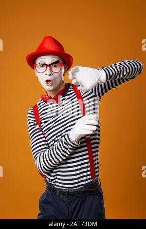 Jeune mime sérieux avec des gants blancs et un chapeau rouge regardant l'appareil photo sur fond orange vide Banque D'Images