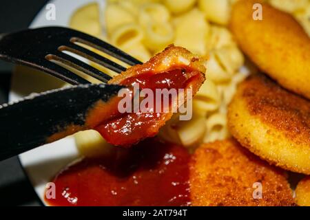 Un morceau de pépites avec ketchup sur une fourchette et une plaque blanche avec des pépites de poulet frits, des pâtes et du ketchup sur un fond sombre. Gros plan Banque D'Images