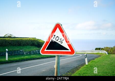 Un triangle rouge indique une pente raide de 10 pour cent dans la route à l'avant. Route vide avec barrières de collision entourées d'herbe verte en arrière-plan. Panneaux de signalisation. Banque D'Images