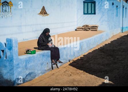 Nubian Village, Assouan, Egypte - Nubian Woman In Traditional Dress Assis Et Counting Money En Face D'Une Maison Bleue Nubienne. Banque D'Images