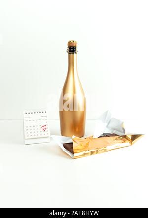 Carte de Saint-Valentin, bouteille de champagne dorée, chocolat dans un wrapper or et calendrier de février avec une date encerclée sur une surface blanche. Vue de face. Banque D'Images
