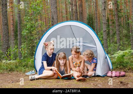 Quatre filles d'âge différent lisant un livre assis ensemble dans le camping de tente dans une forêt d'été pendant les vacances d'été Banque D'Images