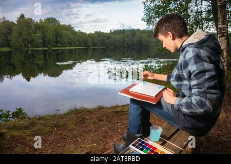 Adolescent garçon assis sur un banc en bois près d'un paysage de peinture de lac forestier avec aquarelles - plein air de peinture concept Banque D'Images