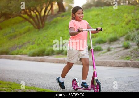 Une petite fille qui monte un scooter rose avec confiance dans une rue bordée d'arbres et d'herbe. Banque D'Images