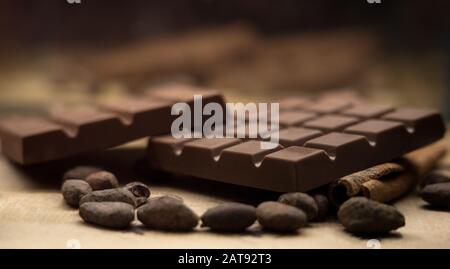 Les barres de chocolat brunes, les fèves de cacao et le bâton de cannelle sur la table sont rapprochés avec un fond flou Banque D'Images