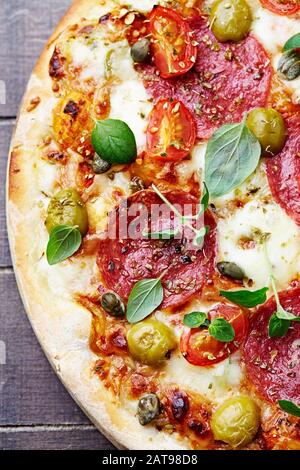 Pizza avec salami, olives vertes, fromage mozzarella, câpres et basilic frais. Fond en bois marron. Vue de dessus. Banque D'Images