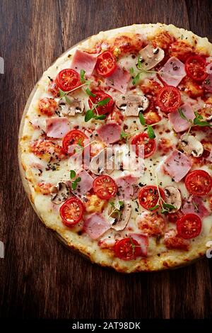 Pizza au jambon, au fromage, aux tomates cerises, aux champignons chamignon et aux herbes fraîches. Fond en bois marron. Vue de dessus. Banque D'Images