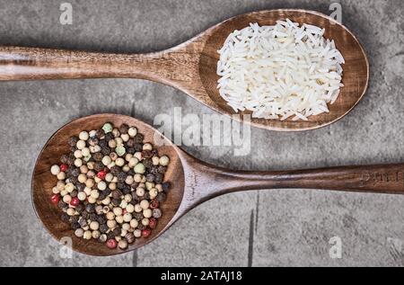 Grains de poivre mélangés et riz non cuit sur deux cuillères brunes et en bois situées sur une surface grise texturée. Vue de dessus et photographie macro-alimentaire. Banque D'Images