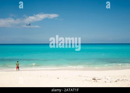 La mer turquoise et le sable blanc de la plage des dauphins à Cancun, Mexique, un homme observe l'horizon, en arrière-plan un parachute ascensionnel avec les touristes Banque D'Images