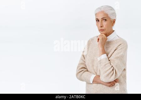 Femme sénior élégante et élégante, élégante et élégante, sénior avec cheveux peignés gris tenue habillée, toucher le menton et regarder l'appareil photo pensive, penser Banque D'Images
