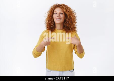 Fière et séduisante jeune fille à tête rouge dans un pull jaune, a été promue, se pointant, souriante enchantée, vantée, plaisante et plaisante racontant à la personne Banque D'Images