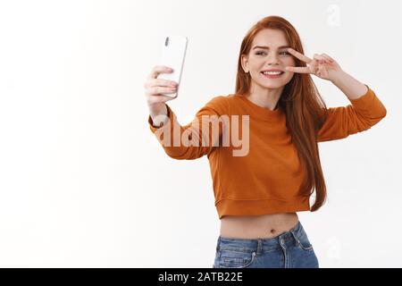 Jolie fille hipster millénaires sans souci avec de longs cheveux rouges prenant selfie à l'aide d'un smartphone blanc, faire signe de paix et sourire, photographier, capturer Banque D'Images