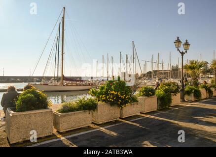 Vue sur le Vieux Port avec des gens et des touristes assis sur les bancs du quai, plantes en pots et bateaux amarrés, Sanremo, Ligurie, Italie Banque D'Images