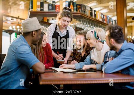Groupe de jeunes amis divers assis ensemble à une table dans un restaurant à la mode de restauration rapide pour commander des repas auprès d'une serveuse Banque D'Images