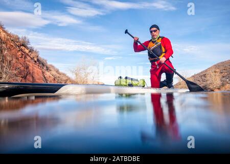 Paddler homme senior dans un costume sec et une veste de vie avec une paddleboard debout sur un lac de montagne en hiver - réservoir Horsetooth à northe Banque D'Images