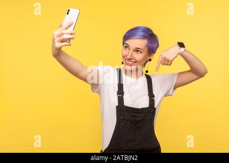 Portrait de gai blogger, fille hipster avec des cheveux courts violettes dans des salopettes tenant le téléphone cellulaire faire un appel vidéo ou faire un blog en ligne et pointage d Banque D'Images