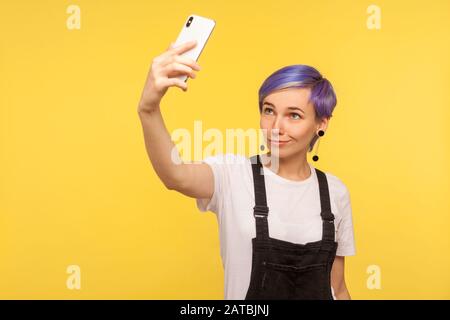 Portrait d'une fille hipster élégante et tendance avec des cheveux violets, une coupe moderne et des salopettes en denim qui tient un smartphone et prend une photo selfie, en faisant vide Banque D'Images
