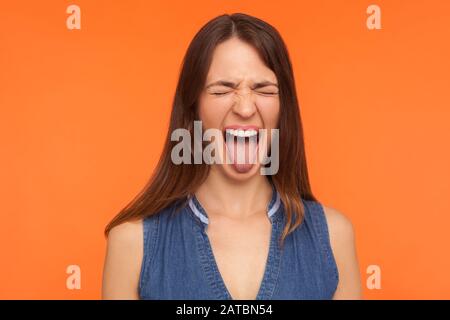 Une femme désobéissante et folle brunette dans des vêtements en denim qui s'enlisent de la langue et grimace aux yeux fermés, qui se moque d'une expression dérisante et méchante. Indo Banque D'Images