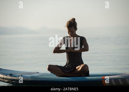 Jeune femme méditant avec ses mains s'est jointe sur sa poitrine, assise sur sup bord flottant sur mer calme matin. Banque D'Images