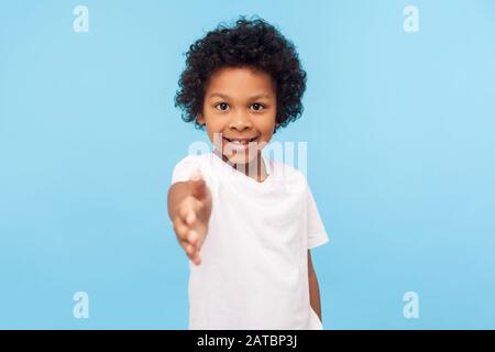 Portrait d'un petit garçon sympathique et gai avec des cheveux bouclés dans un T-shirt blanc donnant la main à la poignée de main et au sourire à l'appareil photo, à la recherche de la confiance créduleuse Banque D'Images