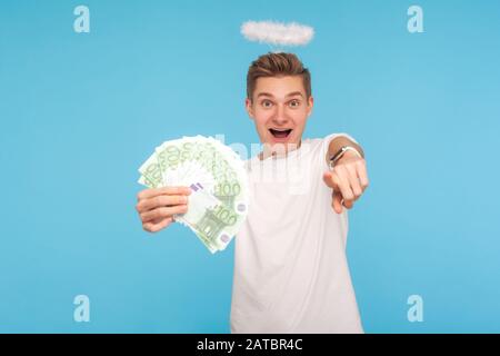 Portrait de l'homme angelique surpris dans le t-shirt et avec halo sur la tête tenant les billets en euros et pointant vers l'appareil photo, exprimant étonnement, choc. Banque D'Images