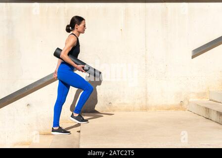installer une femme portant des vêtements de sport, portant un tapis de yoga, marcher sur des marches en béton. Banque D'Images