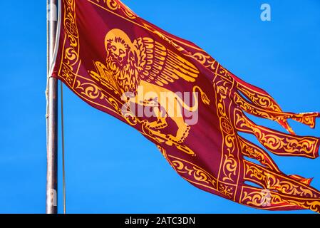 Drapeau de Venise se vengeant dans la rue à Venise, Italie. Lion ailé d'or sur le drapeau rouge, vue rapprochée. Le vieux drapeau vénitien est sur le fond du ciel bleu à summ Banque D'Images