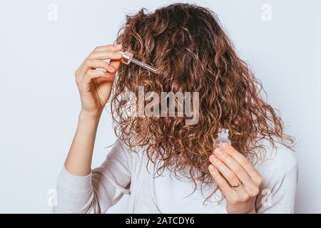 Jeune femme appliquant de l'huile cosmétique à ses beaux cheveux frisés, gros plan sur fond blanc. Banque D'Images