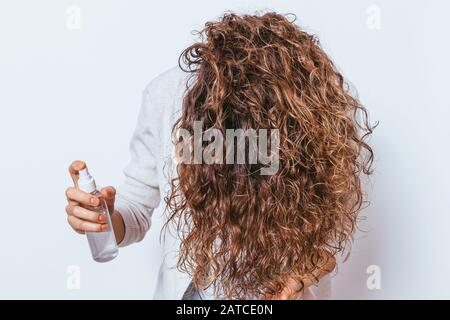 Jeune femme méconnaissable appliquant un spray hydratant à ses beaux cheveux frisés, tenant sa tête vers le bas. Banque D'Images