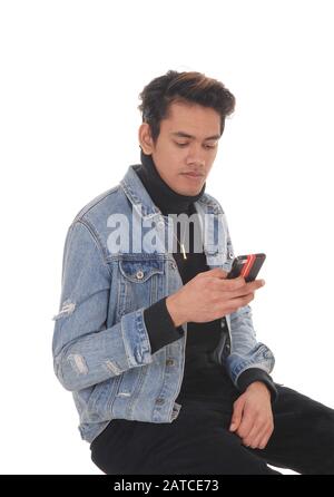 Un jeune homme léger assis dans une veste de Jean et un pantalon noir sur une chaise, regardant son téléphone portable, isolé pour le fond blanc Banque D'Images