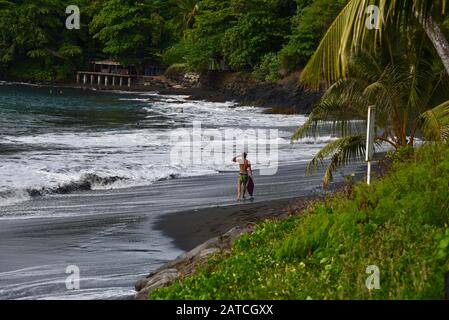 Lone, surfeur masculin, marchant sur une plage de sable noir de l'île de Tahiti. Banque D'Images