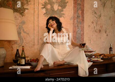 Une femme heureuse en robe de coton blanc avec des noeuds et des volants dans une ambiance festive assise souriante sur une longue table antique, apprécie les plus belles choses de la vie Banque D'Images