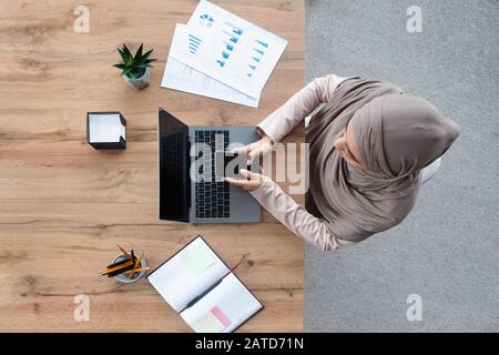Employé Arabe Femme Utilisant Un Smartphone Et Travaillant Sur Un Ordinateur Portable, Vue De Dessus Banque D'Images