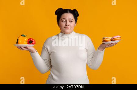 Une fille obèse choisissant entre deux assiettes avec des légumes frais et des beignets Banque D'Images