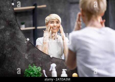 Dame Touchant La Peau Du Visage Regardant Dans Miroir Dans La Salle De Bains Banque D'Images