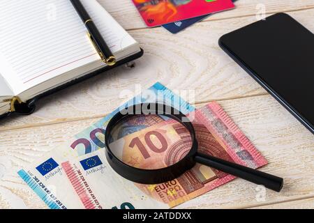 Loupe sur les billets en euros près du stylo, du carnet, des cartes de crédit et du smartphone sur une surface en bois blanc. Authenticité de l'argent et du concept d'affaires. Banque D'Images