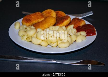 Nuggets de poulet frits et pâtes bouillies avec ketchup sur une plaque blanche sur fond sombre. Gros plan Banque D'Images